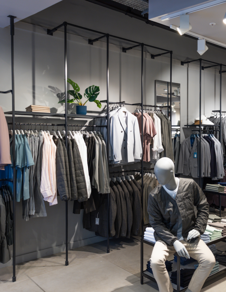 Clothing store display with racks of neutral jackets and a seated mannequin in a quilted gray jacket, plus folded shirts on shelves and a plant on top shelf.